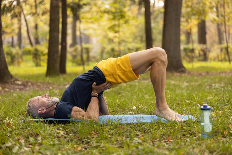 Grizzled bearded male with bottla of water is practicing yoga on mat on grass in nature